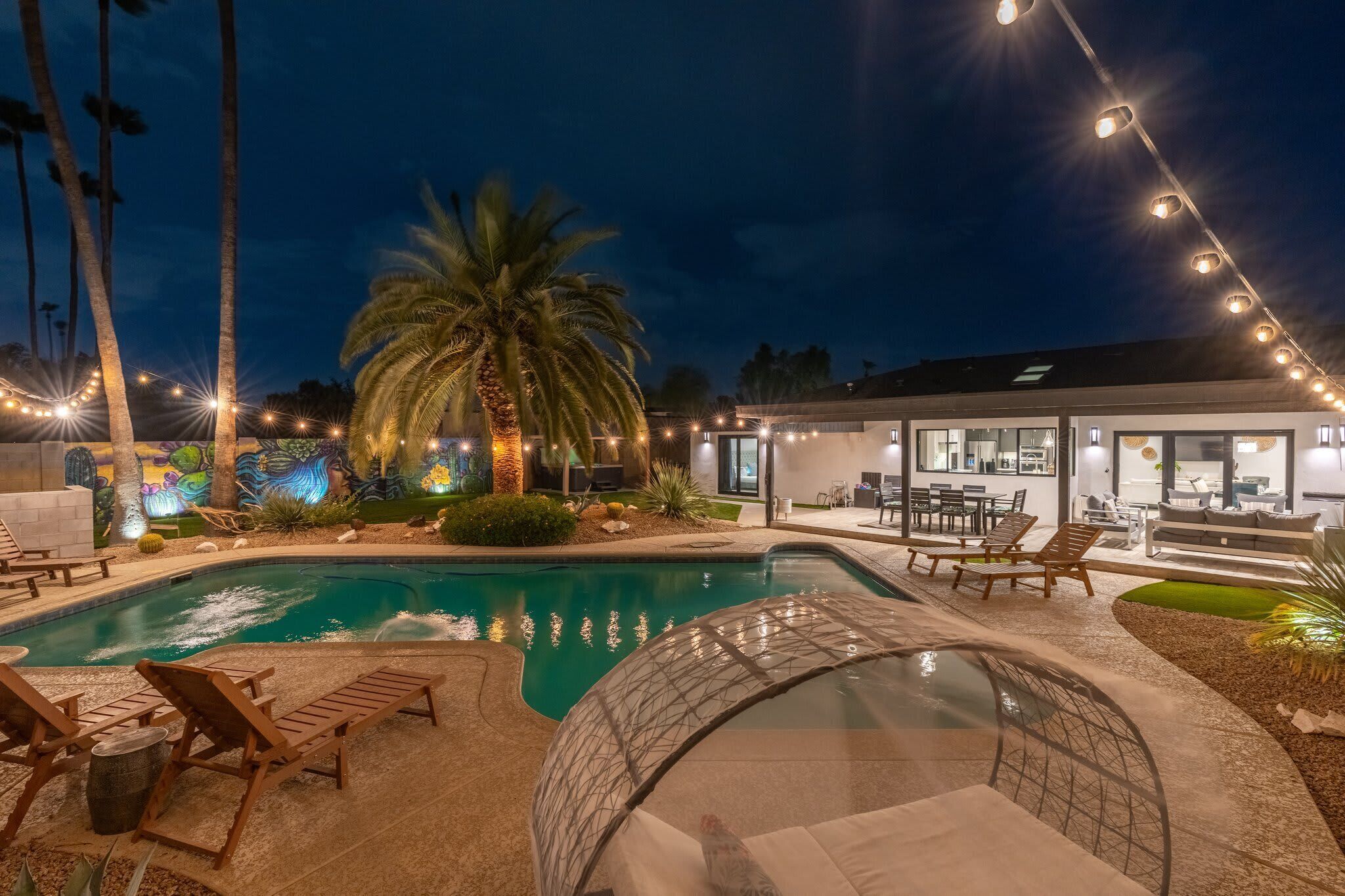 Une piscine avec des chaises longues, un espace de détente couvert et une maison moderne dotée d’un éclairage extérieur.