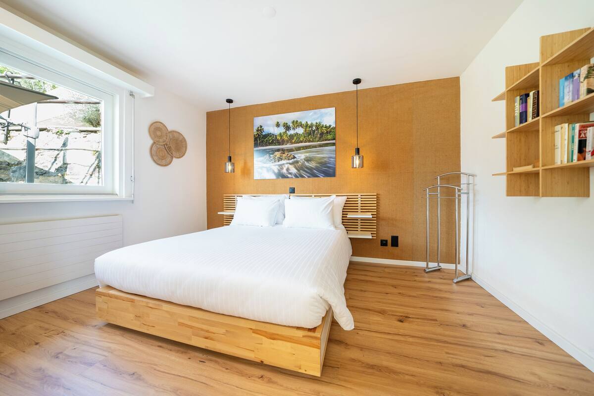 Een moderne slaapkamer met een houten bed, een groot raam, een wandplank met boeken en een schilderij van een tropisch landschap.