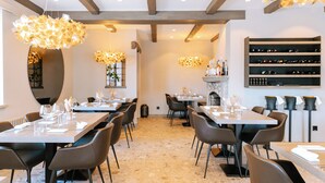 A modern dining area with high ceilings, exposed beams, and pendant lighting. Tables are set with wine glasses and cutlery.