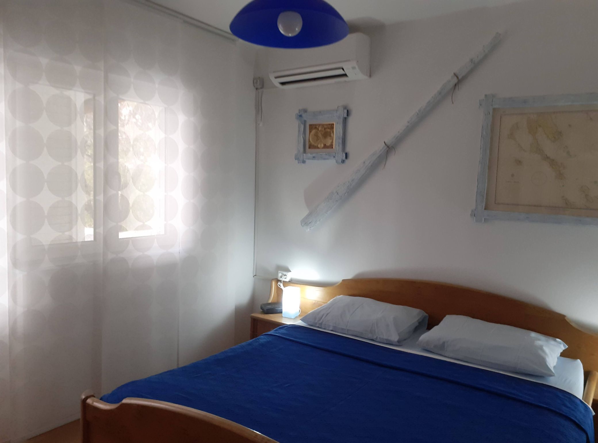 A bedroom with a bed, a nightstand, a lamp, a window with patterned curtains, and a wall-mounted air conditioner.