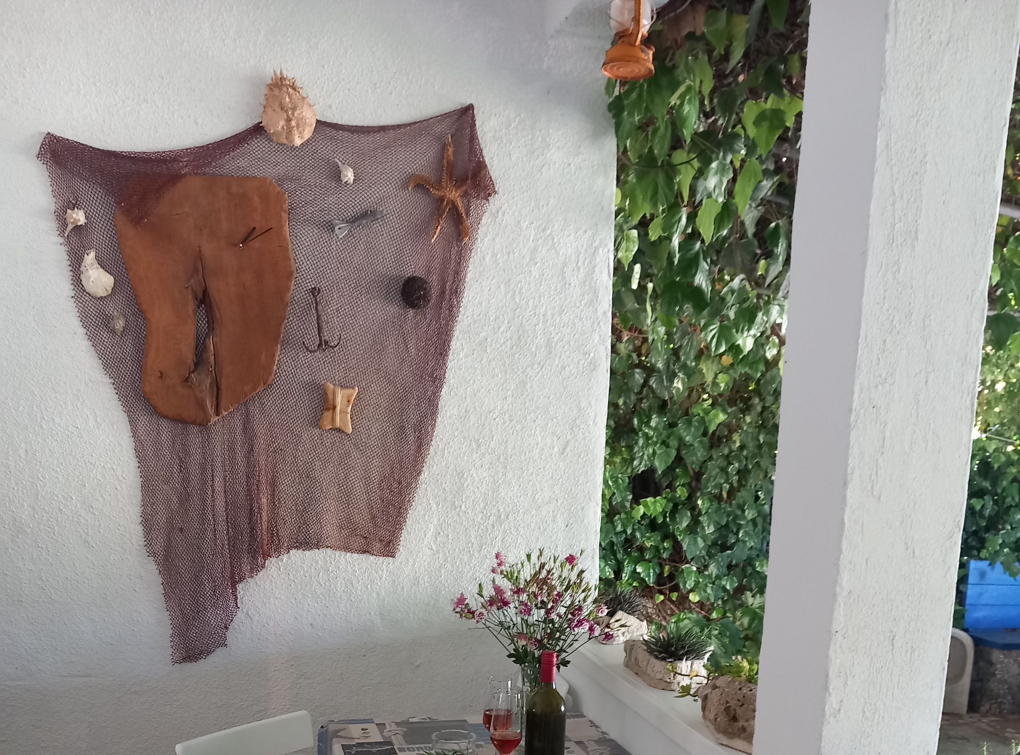 A wall with a woven tapestry, a glass vase with flowers, and a bottle of red wine on a table.