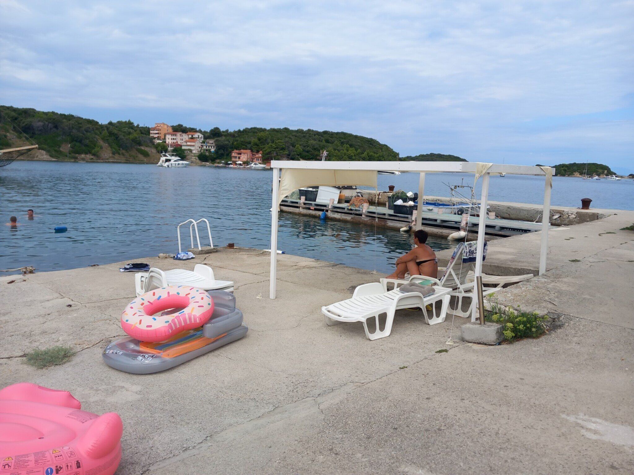 A waterfront area with a covered seating area, inflatable pool, and boats docked in the background.