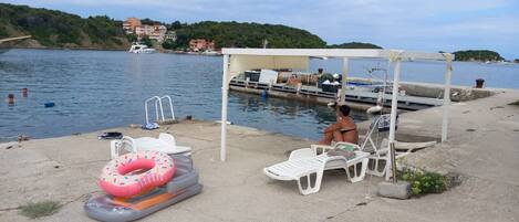 A waterfront area with a covered seating area, inflatable pool, and boats docked in the background.