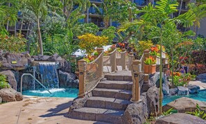 Un área de piscina con cascada, escalones de piedra y plantas tropicales.