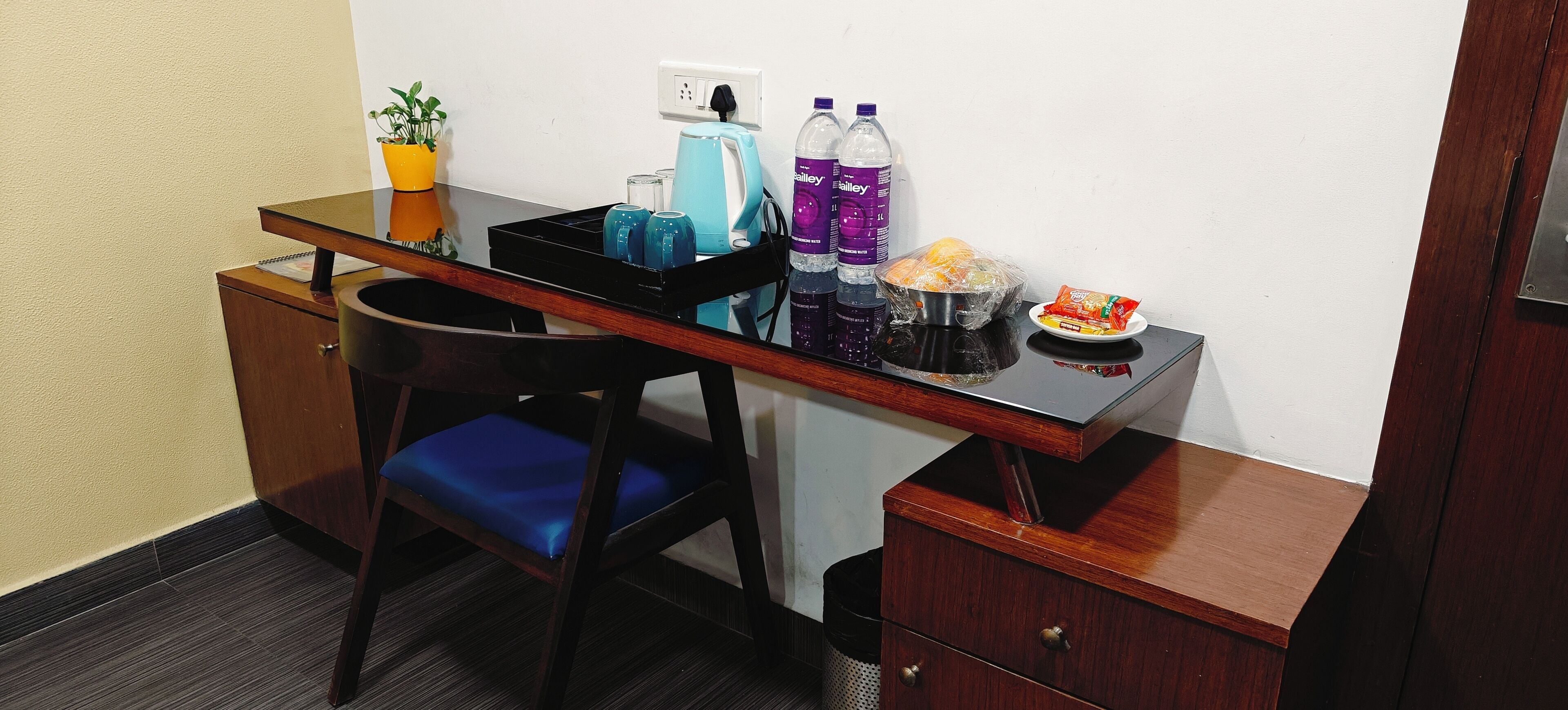 Et kompakt hotelværelse med skrivebord i træ, en blå polstret stol, en lille potteplante, elkedel, flaskevand, en bakke med snacks og en skraldespand.