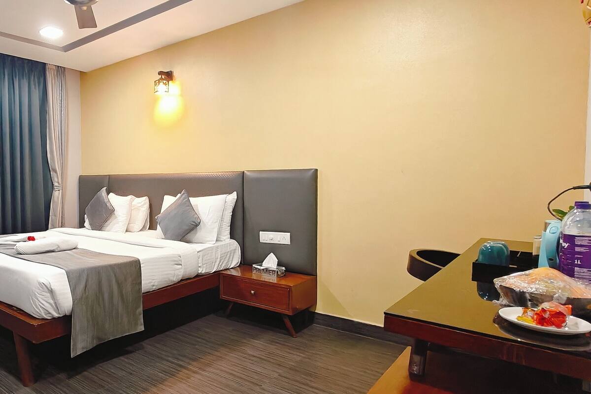 Quarto de hotel com cama, mesas de cabeceira, escrivaninha com cadeira, cortina e luminária fixada na parede.