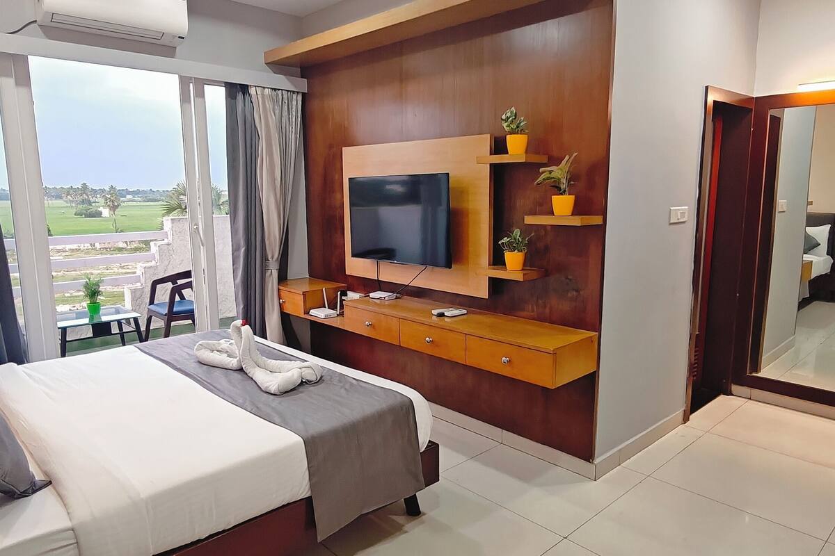 Quarto de hotel com cama, televisão, cómoda de madeira e uma janela grande com cortinas.