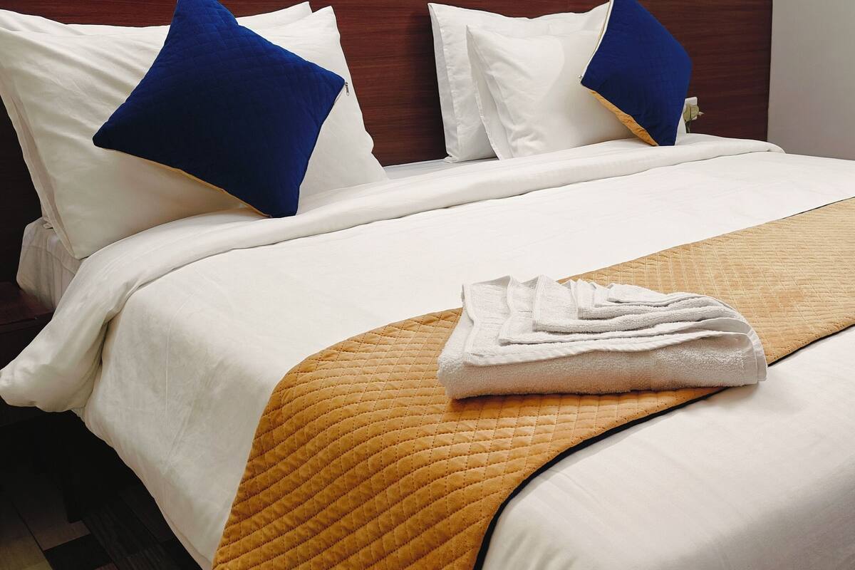 Cama bem arrumada com almofadas brancas e azuis, toalha branca dobrada e manta acolchoada bege.