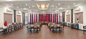 A banquet hall with rows of tables and chairs, air conditioning units, and a stage with curtains. - PGC Chennai Resort & Club By SSS (Ponneri)