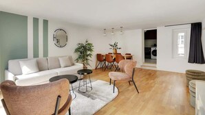 Un salon moderne comprenant un canapé, des fauteuils, une table à manger et une cuisine visible à travers une porte ouverte.