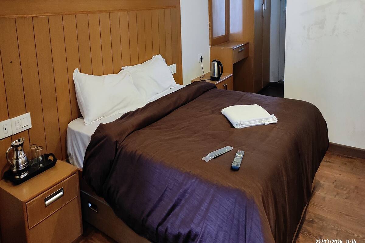 Une chambre d’hôtel avec une tête de lit en bois, un lit avec une literie marron et blanche, une table de chevet avec une bouilloire et des verres, et une télécommande sur le lit.