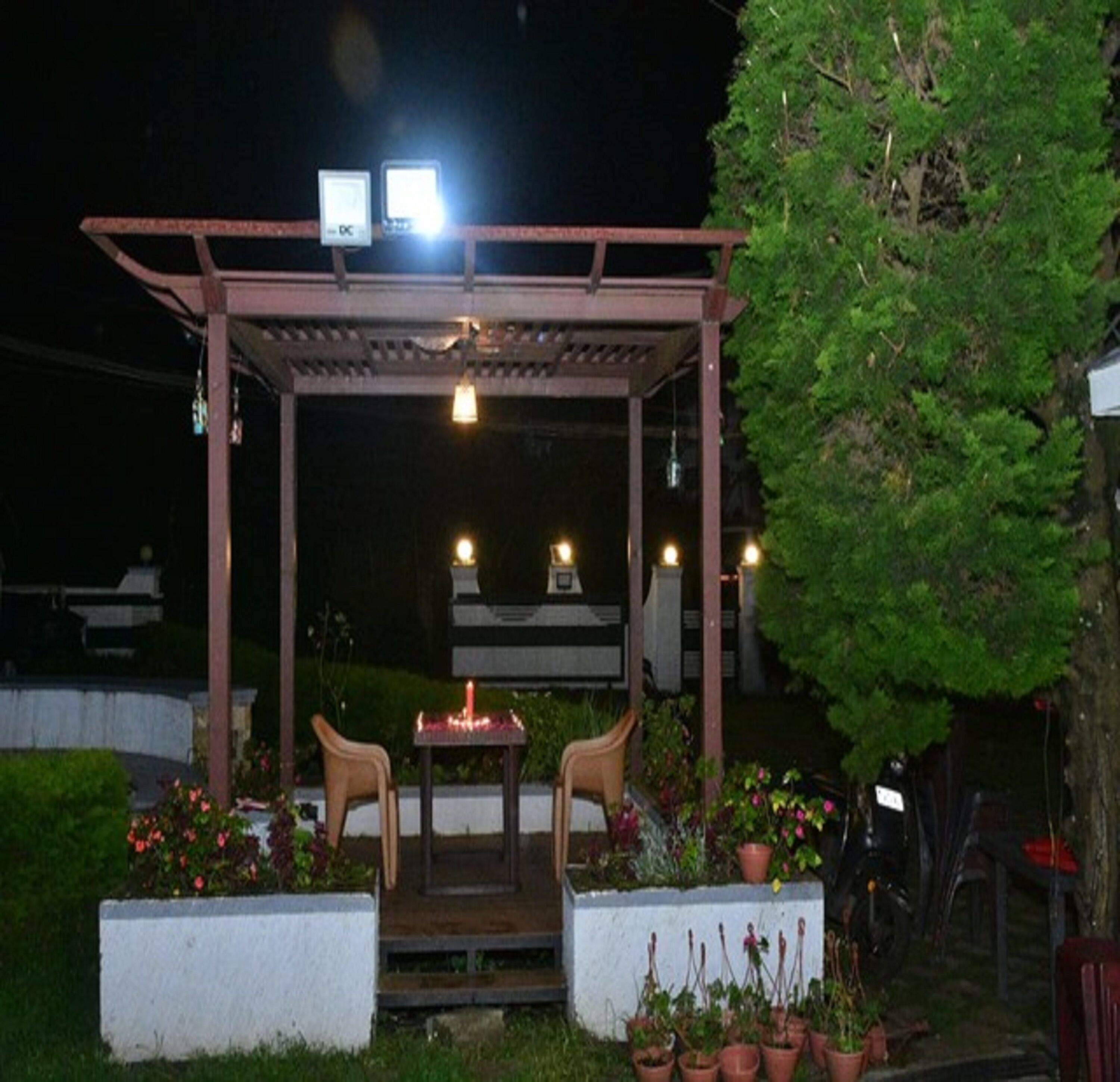 Un espacio de estar al aire libre cubierto con dos sillas y una mesa, rodeado de plantas en macetas y un árbol.