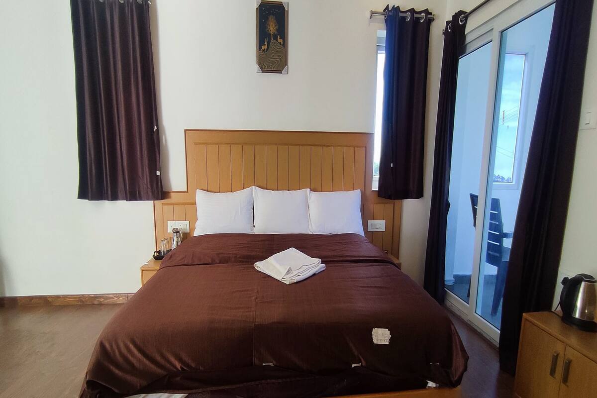 Une chambre d’hôtel avec un lit en bois, des oreillers blancs, une couvre-lit marron, une table de chevet, une petite table avec une lampe, un tableau au mur et une vue sur l’extérieur par une porte coulissante.