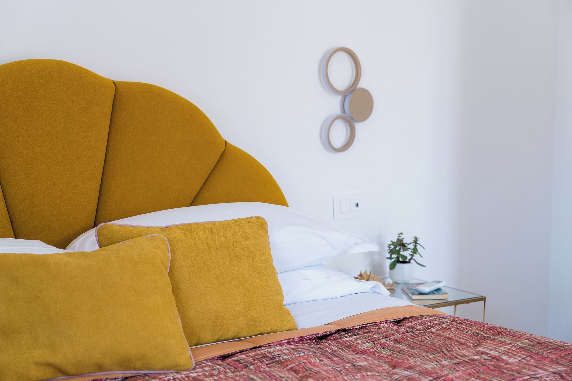 A neatly made bed with a mustard-colored headboard, white bedding, and a patterned red blanket. There is a small bedside table with a plant and a book.