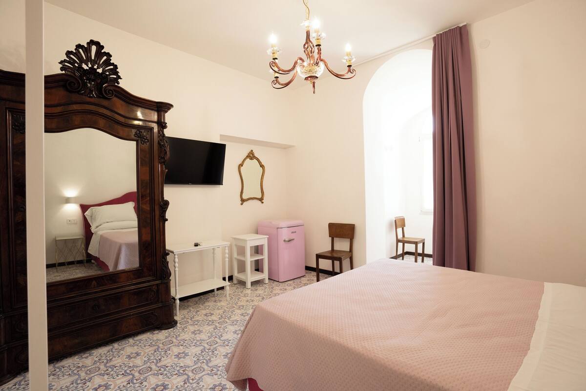 A bedroom with a large bed, a dresser, a vanity, a mirror, a television on the wall, and a window with curtains.