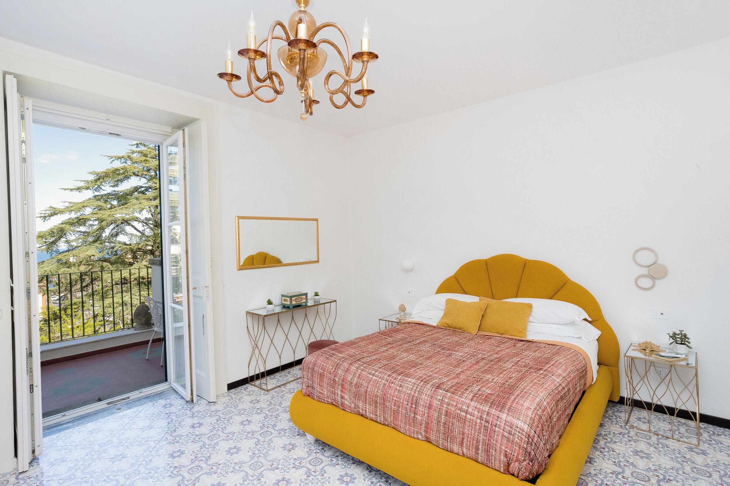 A bedroom with a bed, a chandelier, a desk, and a balcony with a view of trees.