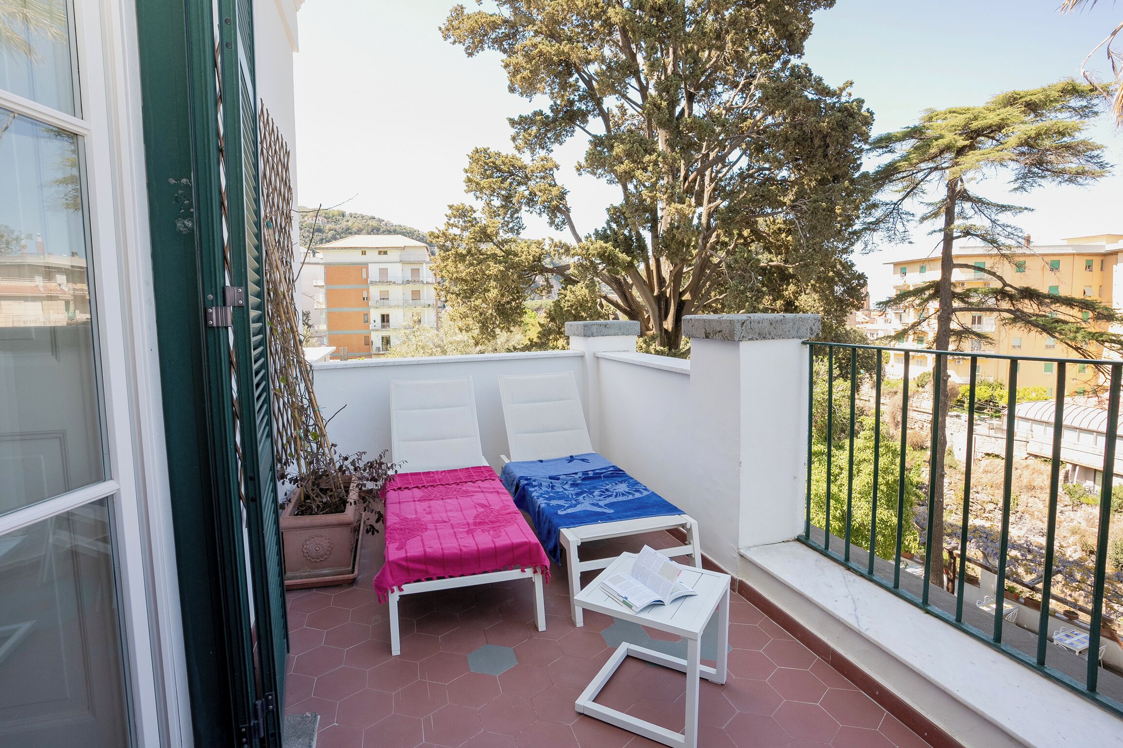 A balcony with two white chairs, one with a pink and one with a blue cover, and a small table with a book.