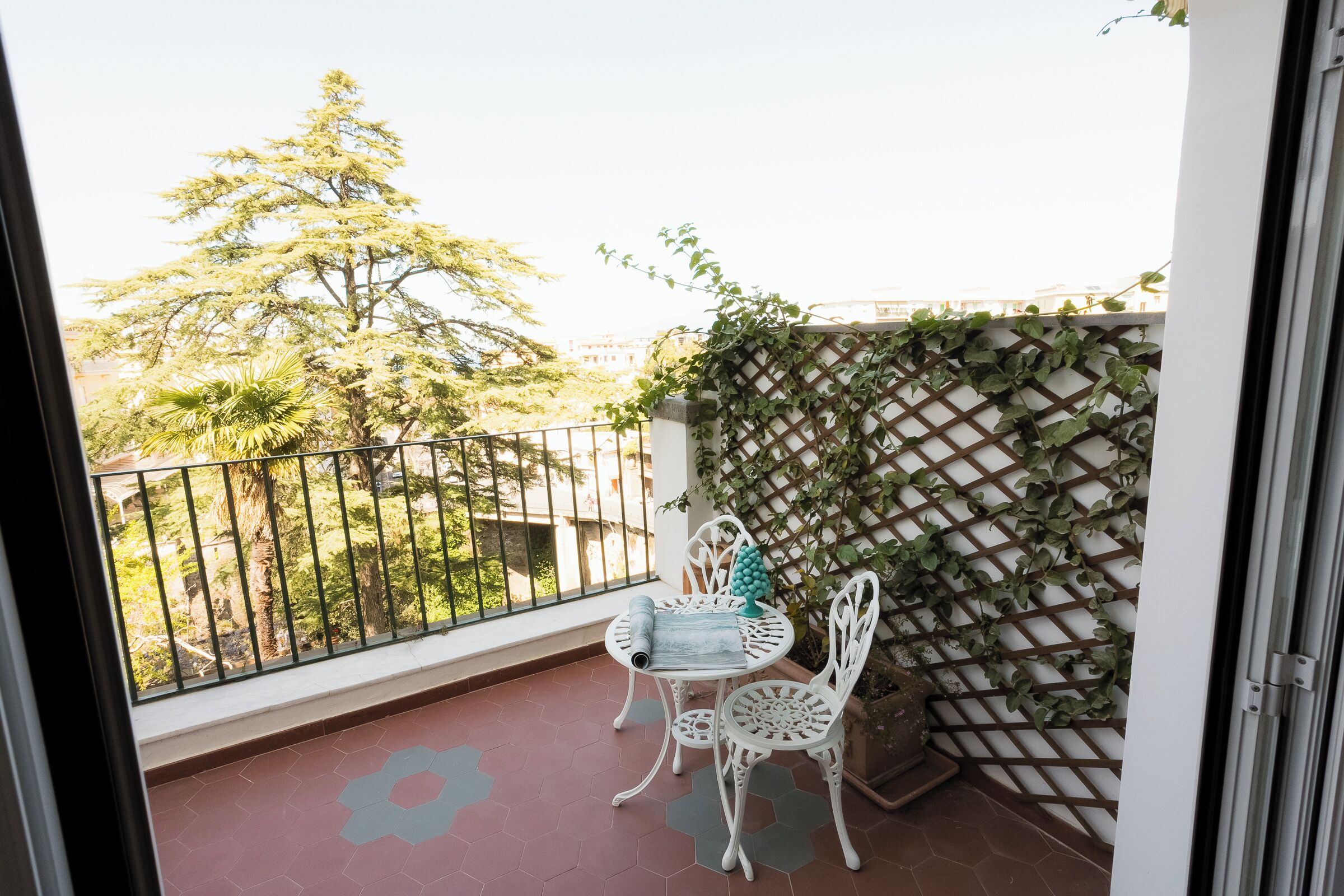 A balcony with a white metal table and chairs, a patterned floor, and a view of trees and buildings.