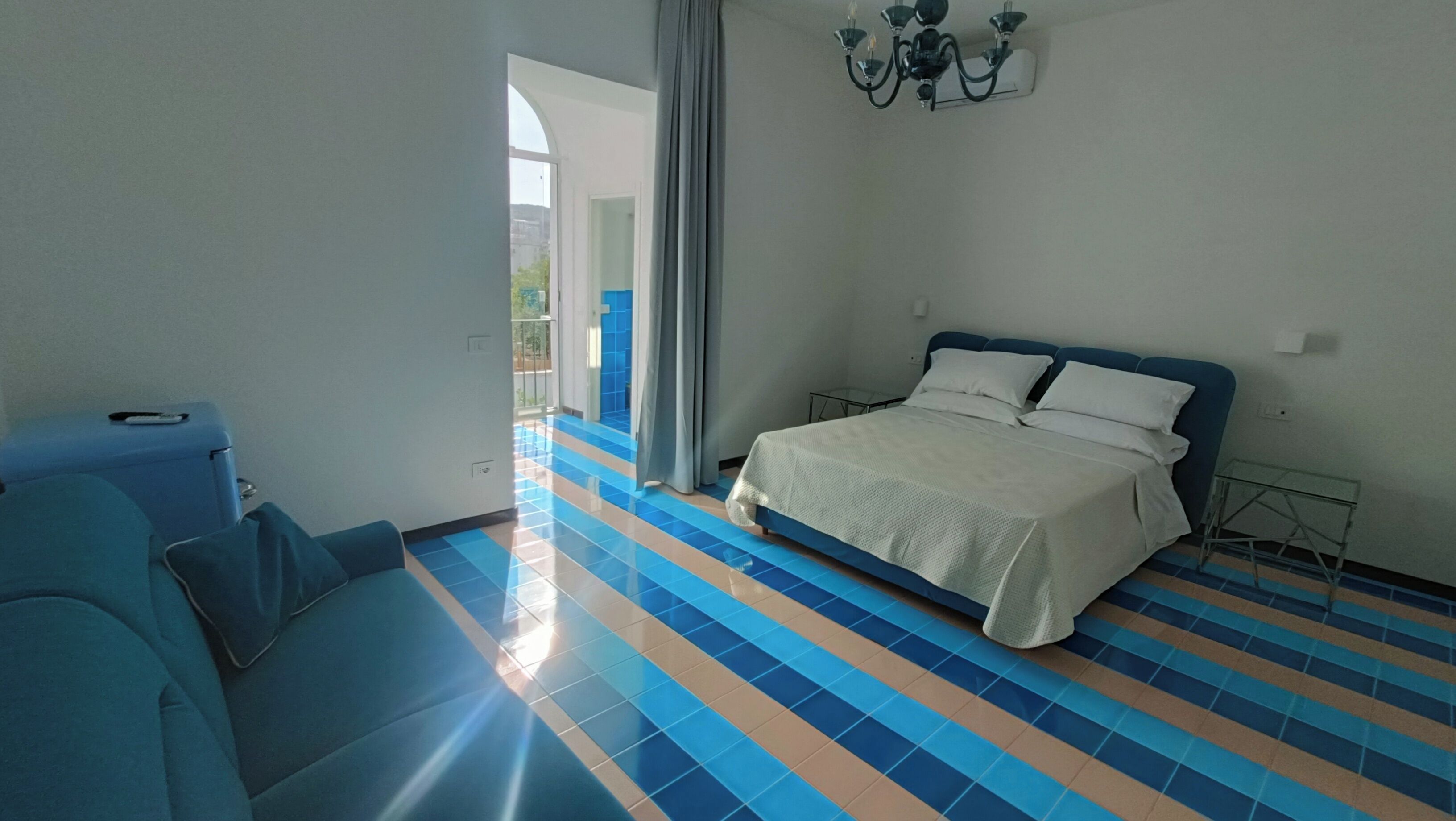 A hotel room with a bed, a blue armchair, a suitcase, and a balcony with blue and white tiles.