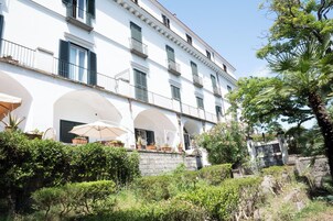 A white building with multiple balconies and arched windows. There are potted plants and outdoor furniture visible.