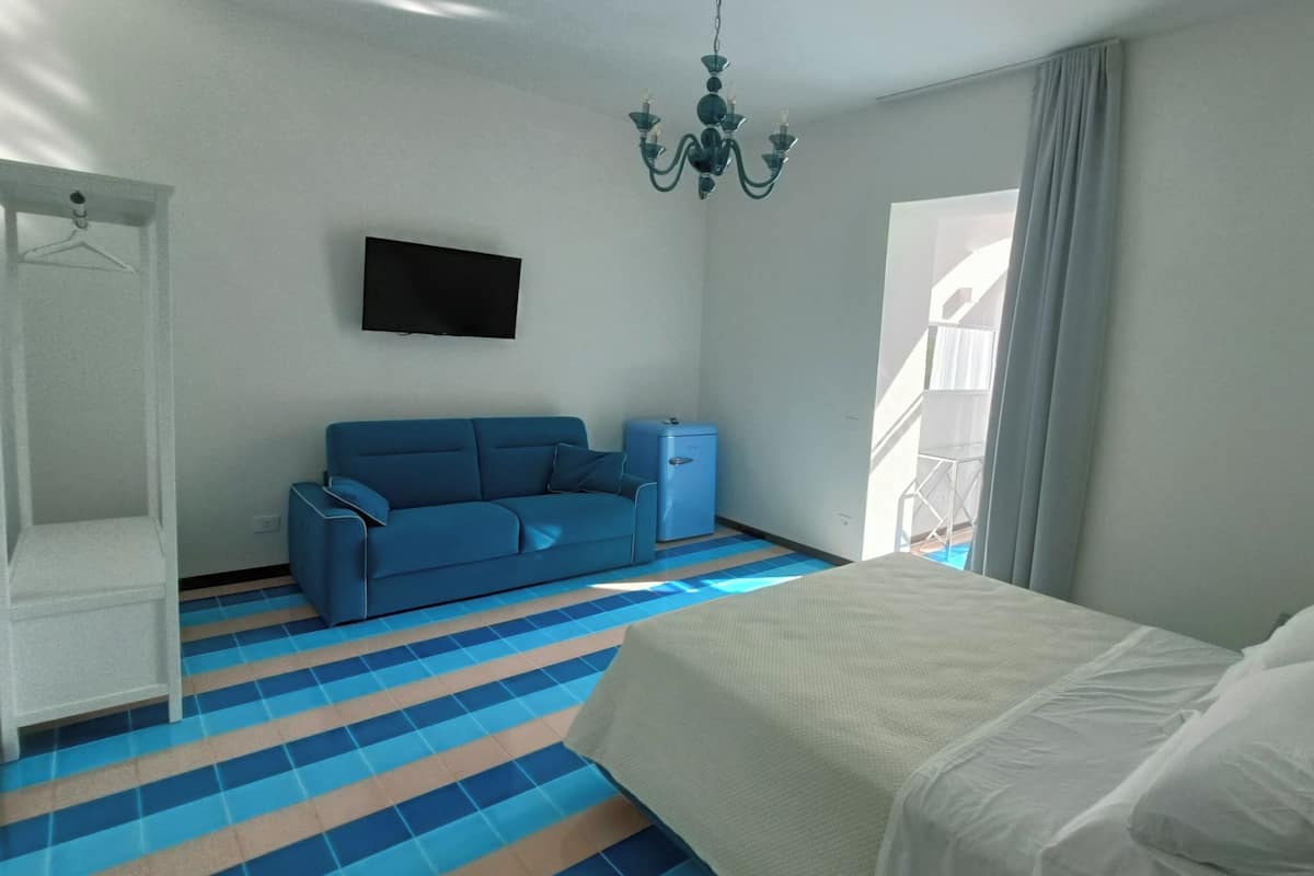 A bedroom with a bed, a blue sofa, a small cabinet, and a TV on the wall.