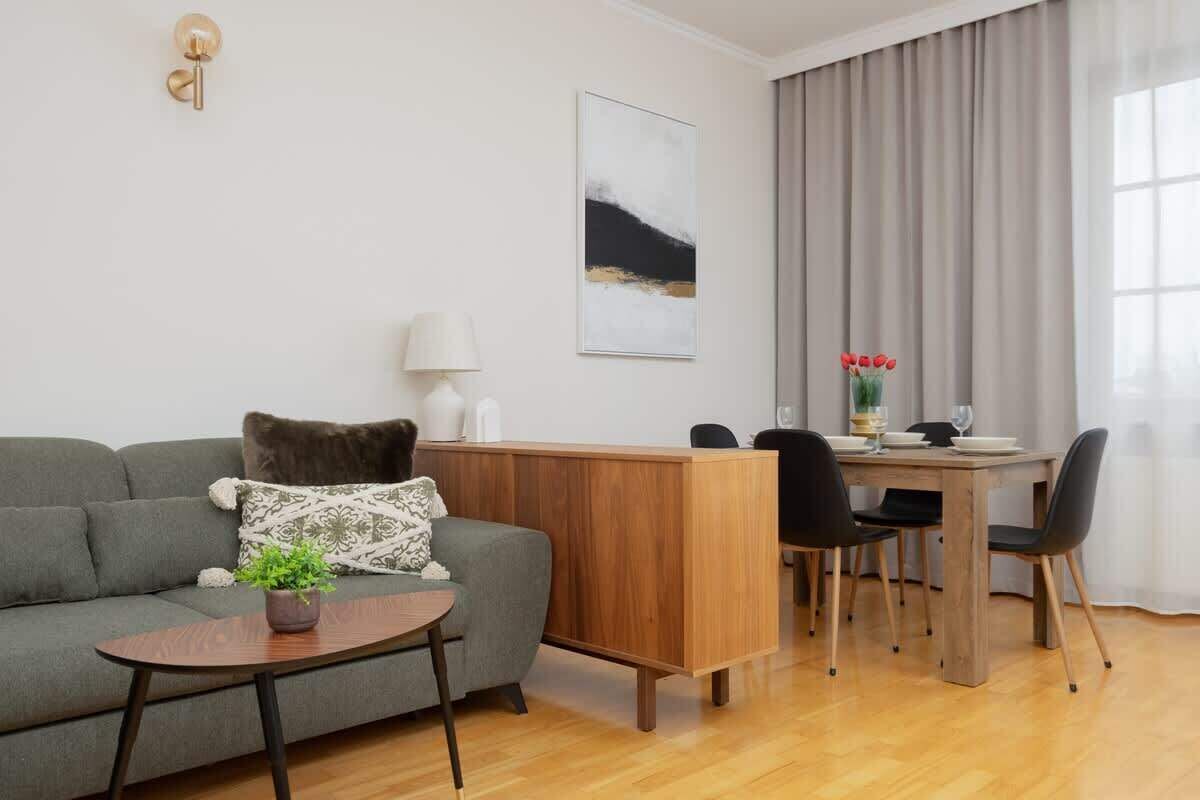 A modern living room with a grey sofa, a wooden coffee table, a dining area with a wooden table and chairs, and a large window with curtains.