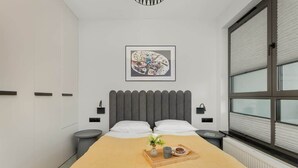 A modern bedroom with a large bed, a bedside table with a tray, a gray headboard, a framed artwork, and a window with blinds. - Modern Apartment with Balcony | Parking, Close to the River (Gdansk)