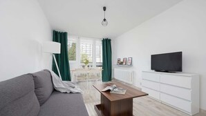 A modern living room with a grey sofa, a wooden coffee table, a white TV stand, and a view of the outside through large windows. - Apartment with 2 Bedrooms in Wola | Workspace (Warszawa)