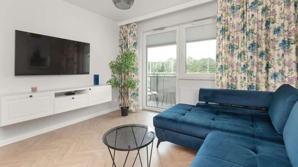 Ein modernes Wohnzimmer mit einem blauen Sofa, einem an der weißen Wand befestigten Flachbildfernseher, einem Glastisch und einer Topfpflanze.