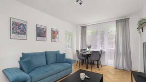 A modern living room with a blue sofa, a black coffee table, and a dining area with a table and chairs. There are two framed artworks on the wall and a window with curtains. - Traditional Apartment with Balcony | Close to the Park (Poznan)