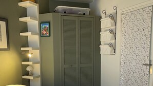 Un lit bien fait, avec des serviettes pliées et une bouteille à côté, une armoire avec des portes coulissantes, un porte-serviettes fixé au mur et une porte ornée d’un motif de feuilles.