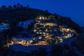 A hillside property with multiple buildings, including a main house with a large patio and a smaller structure with a thatched roof.