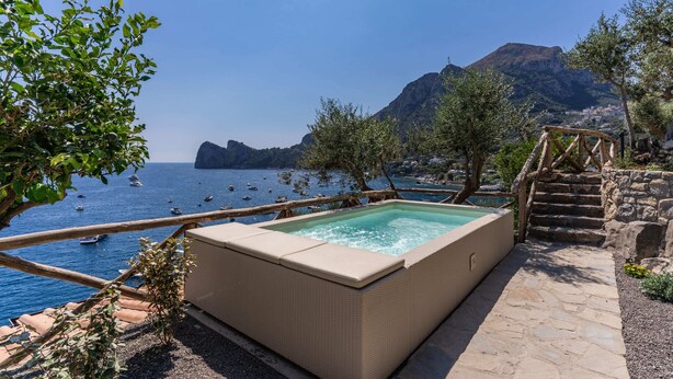 A rectangular hot tub with a stone pathway leading to it, overlooking a body of water with boats and a mountainous backdrop.