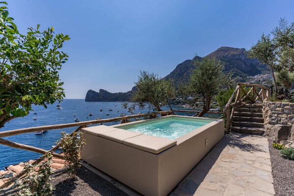 A rectangular hot tub with a stone pathway leading to it, overlooking a body of water with boats and a mountainous backdrop.