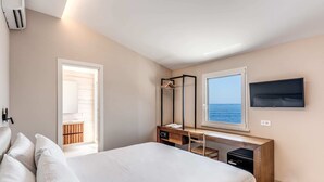 A bedroom with a bed, a desk with a chair, a TV, and a window with a view of the sea.