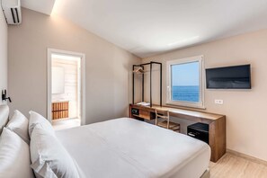 A bedroom with a bed, a desk with a chair, a TV, and a window with a view of the sea.