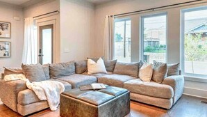 Ein modernes Wohnzimmer mit einem Ecksofa, Pouf und großen Fenstern, die für natürliches Licht sorgen.