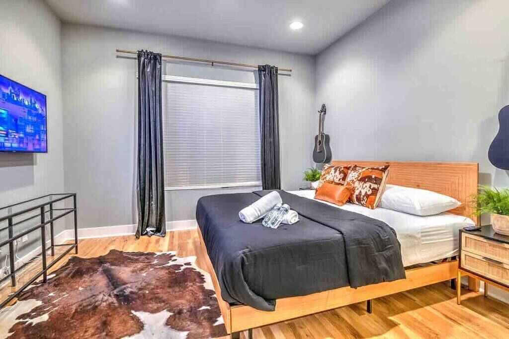 Un dormitorio moderno con una cama de madera, un tapete de cuero de vaca, una guitarra y un televisor en la pared.