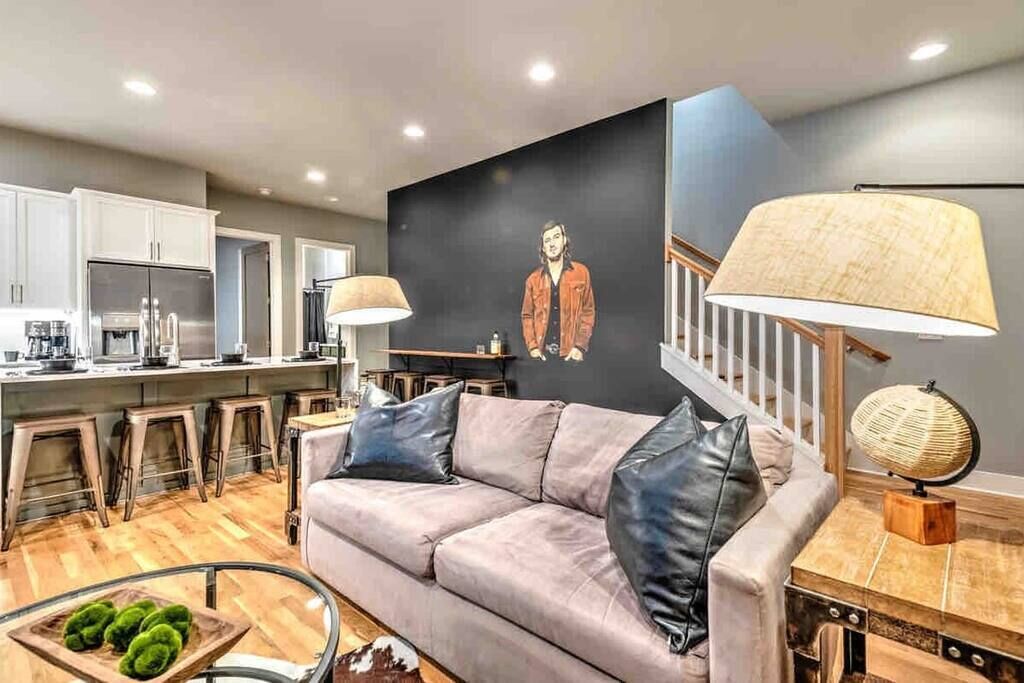 A modern kitchen with white cabinets and stainless steel appliances, a grey sofa with black cushions, and a staircase with a wooden railing.