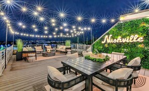 Eine Dachterrasse mit Lichterketten, einem Tisch mit Stühlen und einer Wand mit Pflanzen und einem Neonzeichen.