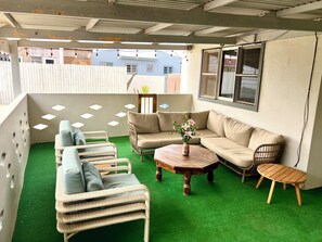 Un área de estar al aire libre cubierta, con mobiliario de mimbre, piso de césped artificial verde y una mesa de madera.