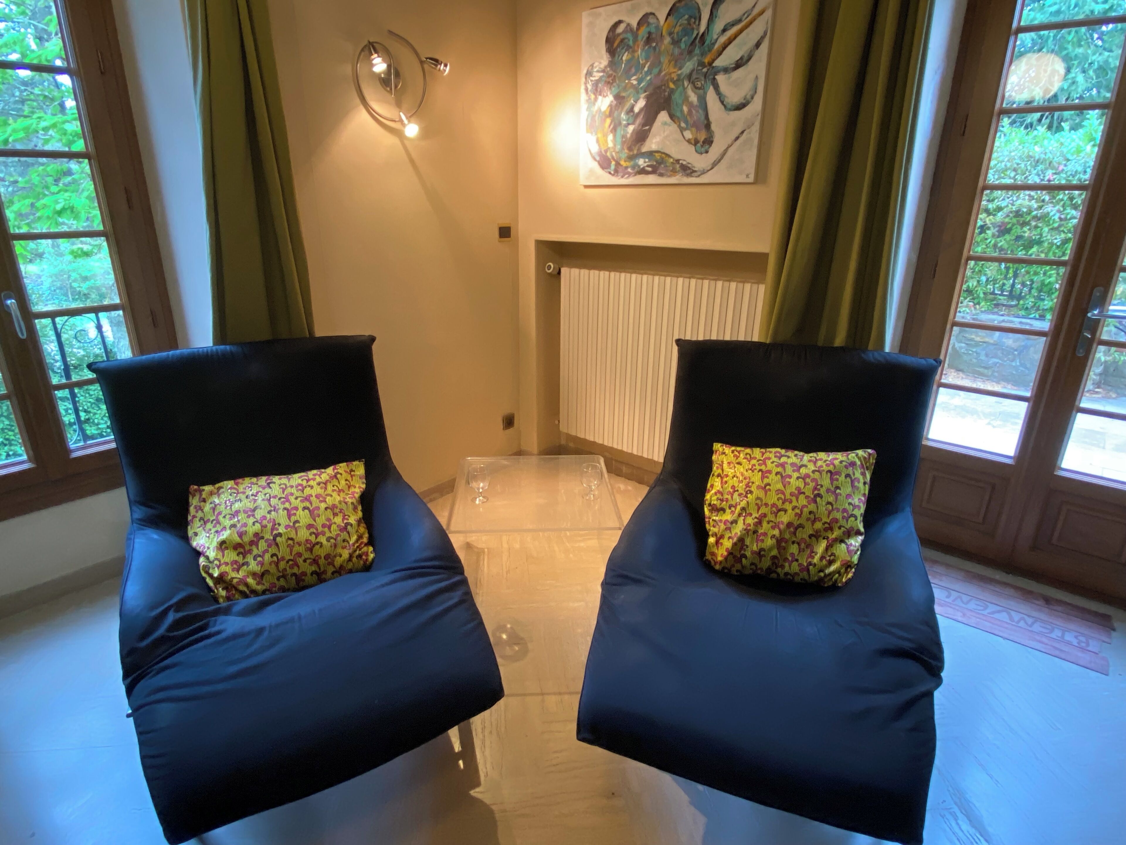 A room with two black chairs with patterned cushions, a glass table, a painting on the wall, and a view of greenery through the windows.