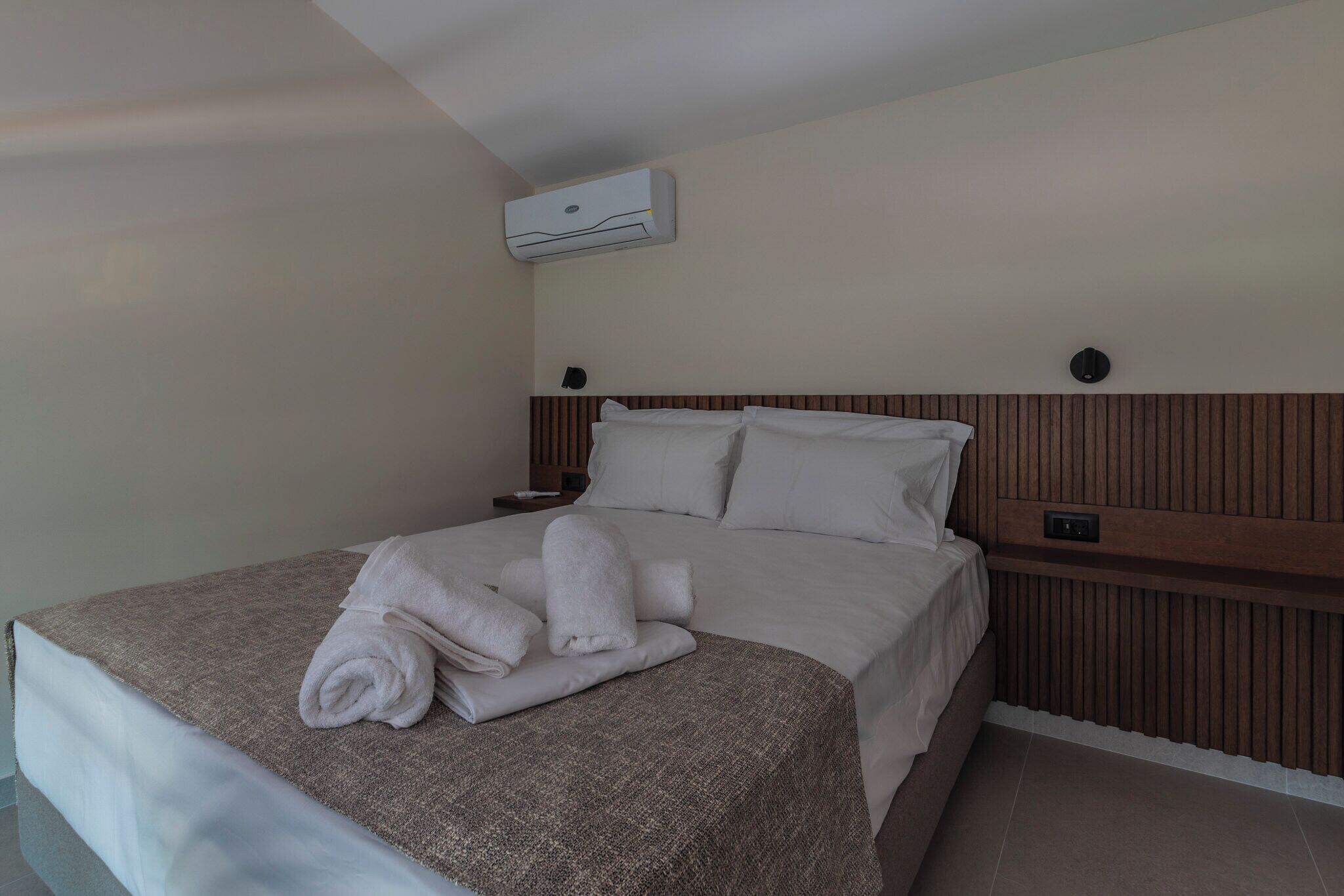 A neatly made bed with a brown blanket and rolled towels, a wooden headboard, and an air conditioning unit on the wall.
