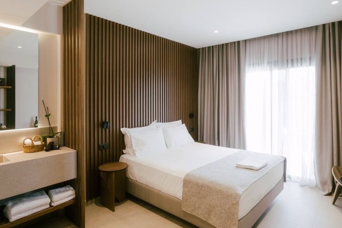 A modern hotel room with a large bed, a bedside table, a vanity with a mirror, and a window with curtains.