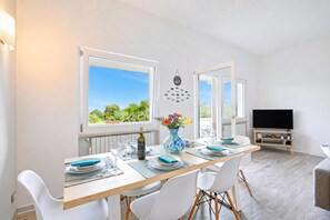 A dining area with a table set for four, a vase with flowers, and a television in the background. - Villa Ulivi private Pool & Lake View (San Felice del Benaco)