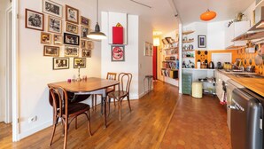 Een eetruimte met een houten tafel en stoelen, een keuken met een koelkast en fornuis, en een gang met ingelijste foto's aan de muur.