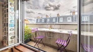 Een raam met uitzicht op een balkon, twee paarse stoelen en een kleine tafel.
