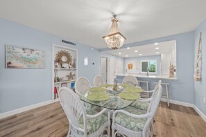 A dining area with a glass table surrounded by white wicker chairs, a chandelier above, and a kitchen in the background. - Loggerhead Cay 242: Beautiful Beachside Condo! (Sanibel)
