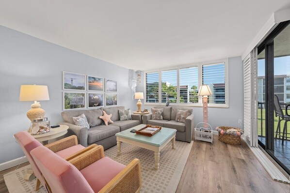 A living room with a grey sofa, a pink armchair, a coffee table, and a lamp. There are framed pictures on the wall and a window with blinds. - Loggerhead Cay 242: Beautiful Beachside Condo! (Sanibel)