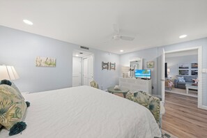 A bedroom with a large bed, a TV, a dresser, and a door leading to a living area. - Loggerhead Cay 242: Beautiful Beachside Condo! (Sanibel)
